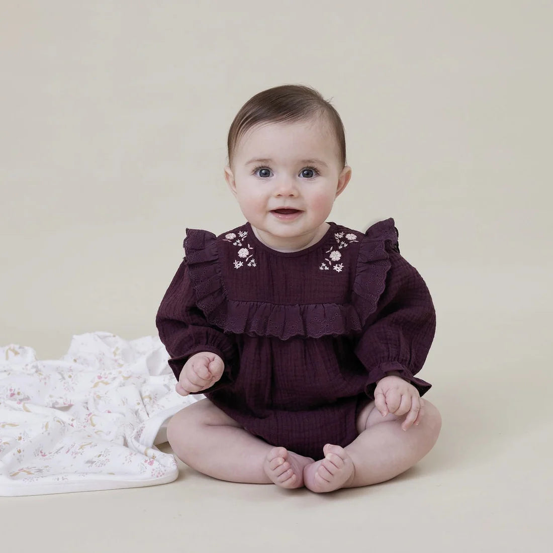 Baby wearing a dark purple outfit with ruffles and floral embroidery on a beige background