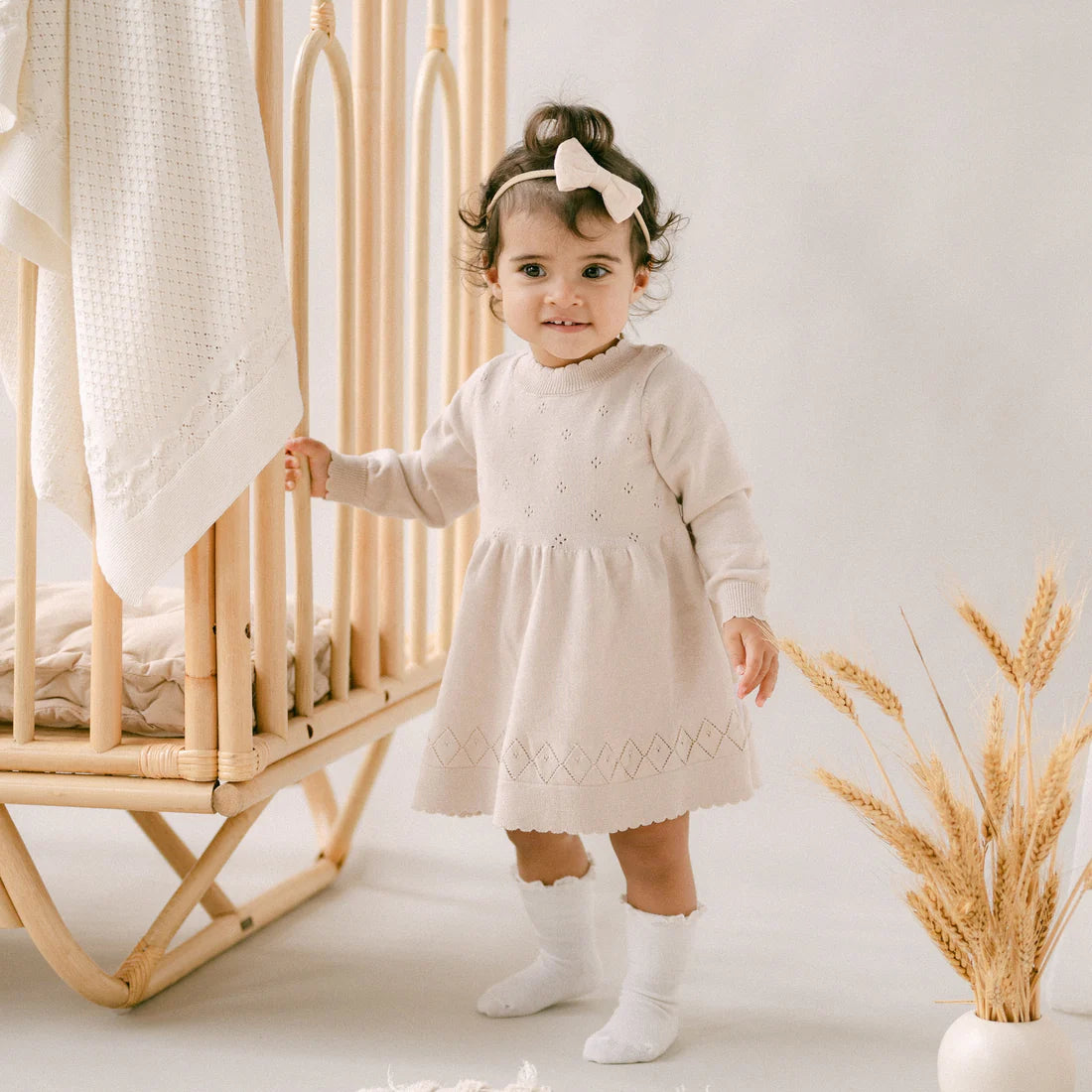 Child in a light dress standing next to a crib with a neutral background