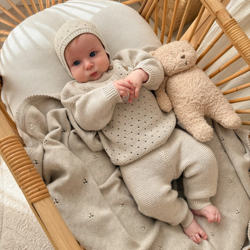 Baby in a knitted outfit sitting in a crib with a teddy bear