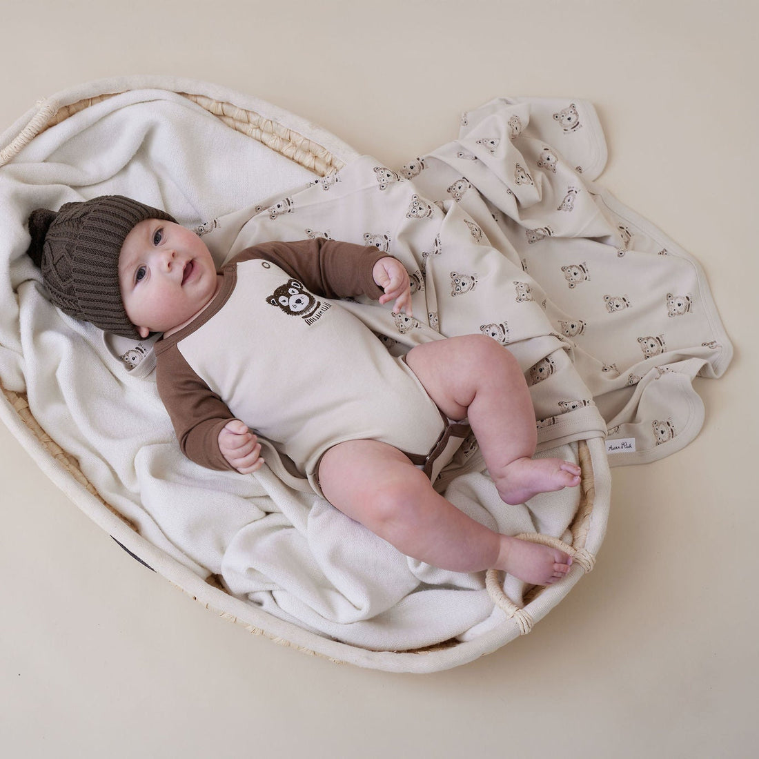 Baby in a brown hat and romper lying on a white blanket with a soft focus background