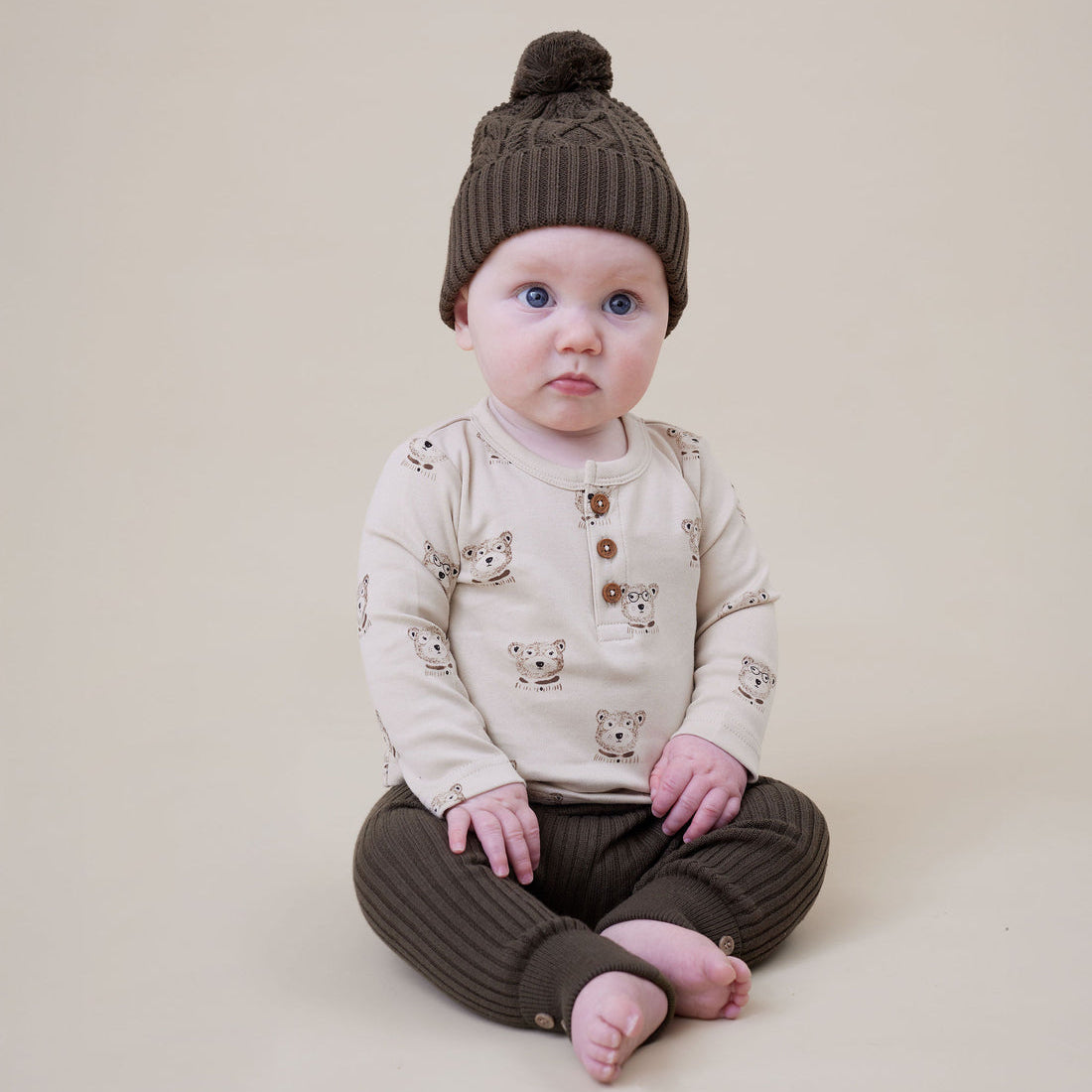 Baby wearing a brown knit hat and matching pants sitting on a beige background