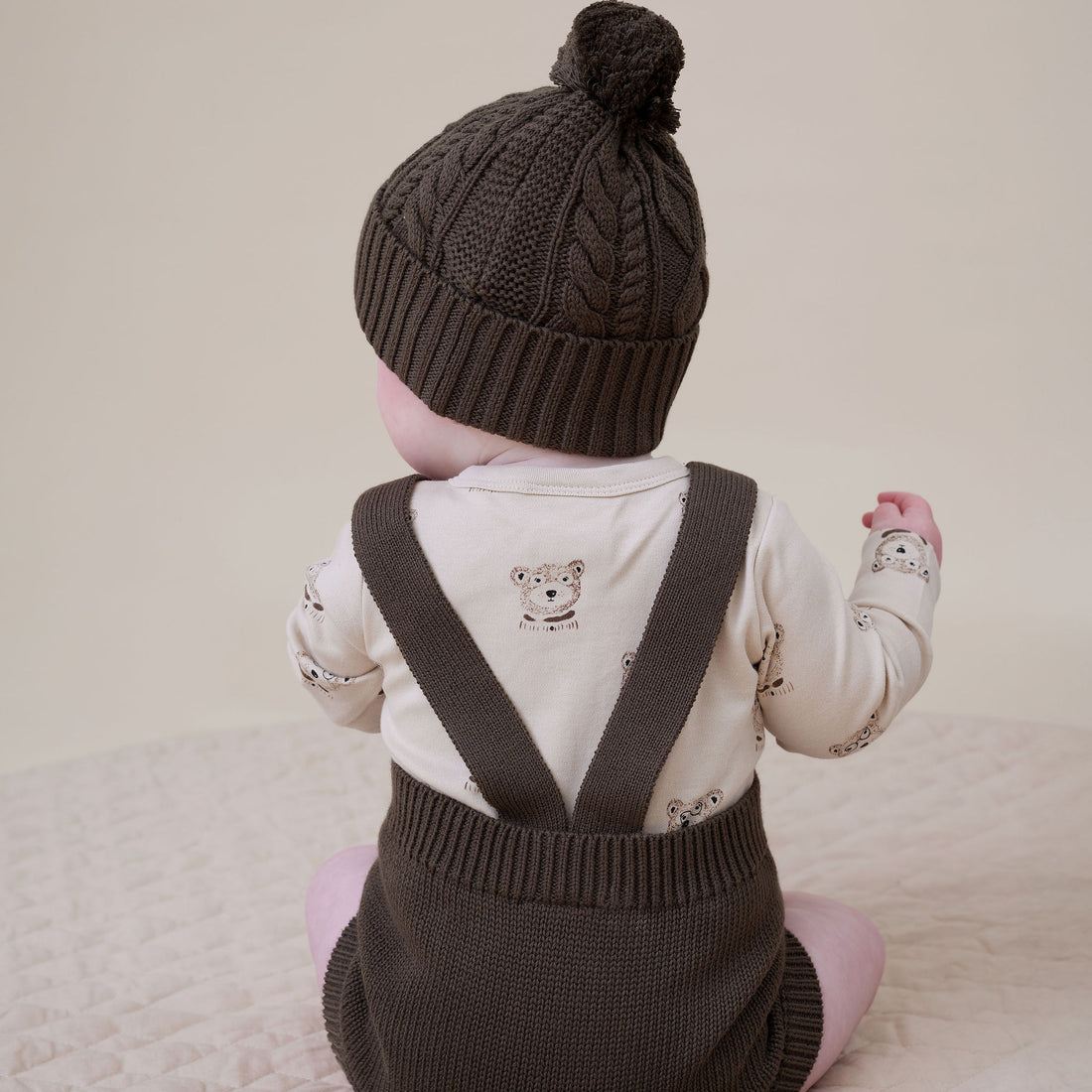 Child wearing a brown knitted hat and matching suspenders on a light background
