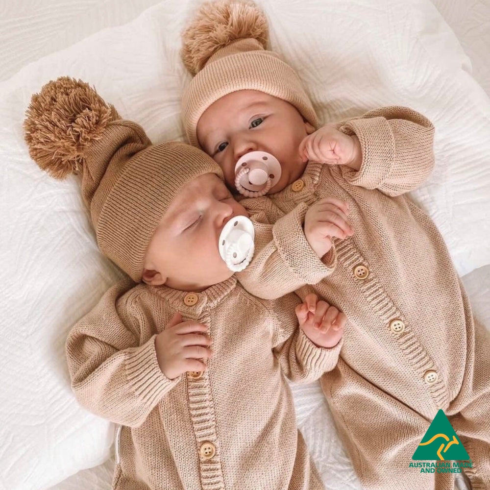 Two babies in matching beige outfits with pom-pom hats lying on a white blanket with round dummies in their mouths