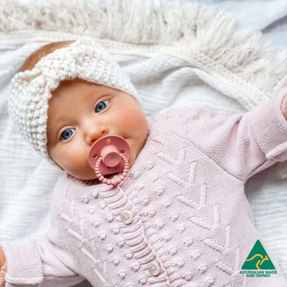 Baby in a pink textured outfit with a white hood and a pink dummy, lying on a soft surface.