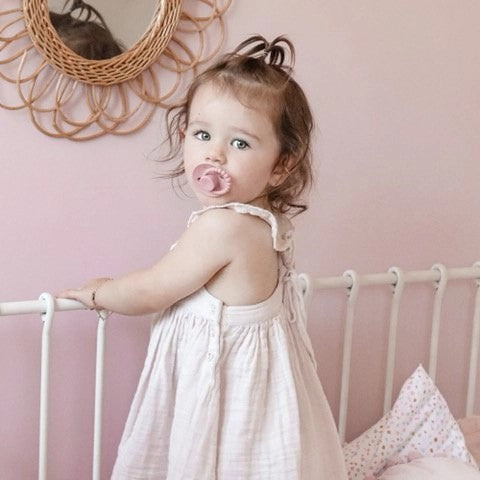 Child in a white dress with a pink pacifier in a room with pink walls and a decorative mirror.
