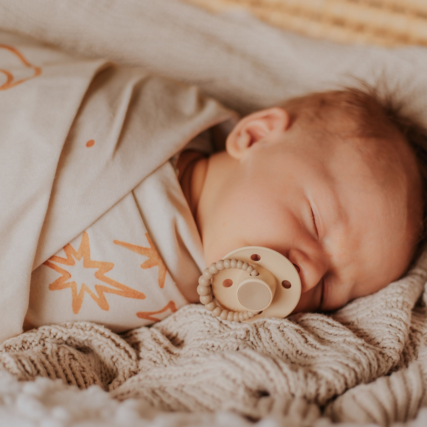 Newborn baby sleeping peacefully with a beige pacifier, wrapped in soft blankets.