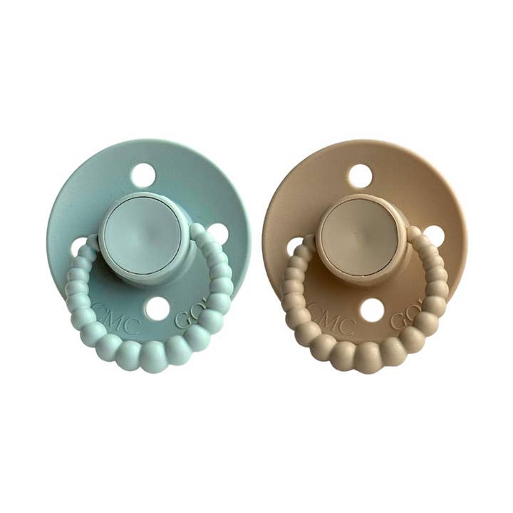 Two pacifiers, one light blue and one beige, with beaded designs on a white background.