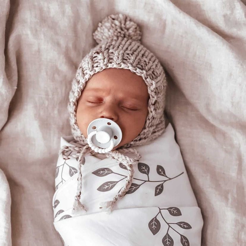 Newborn baby swaddled in a blanket with leaf patterns, wearing a knitted hat and holding a pacifier.