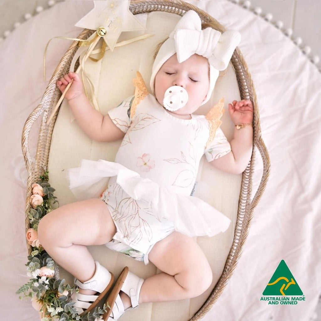 Baby in a white outfit with a pacifier, lying in a decorative basket with flowers, on a light background.