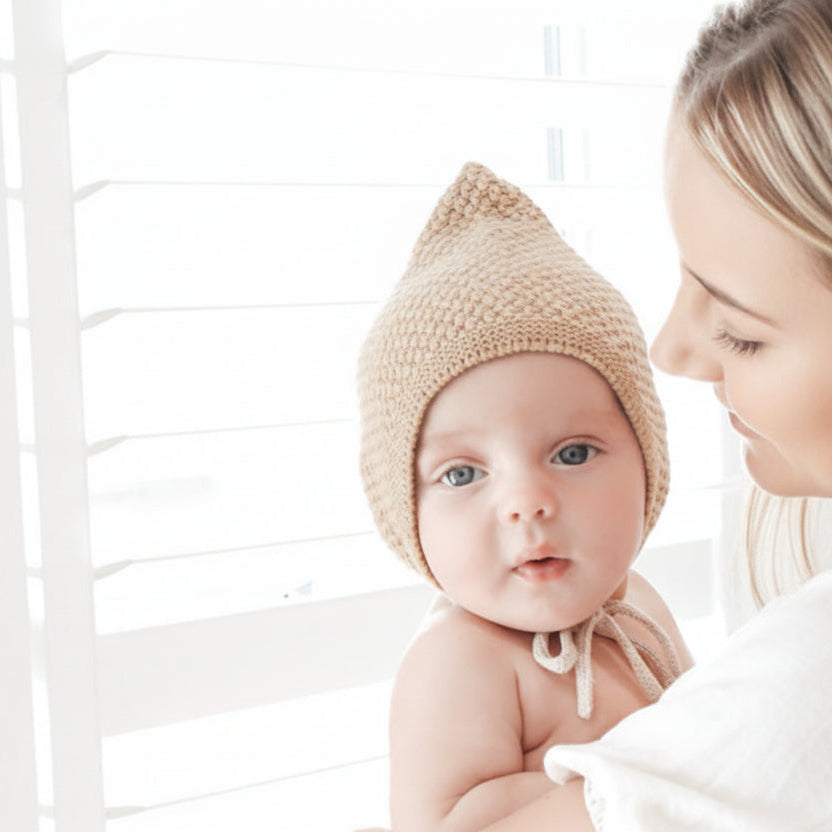 Baby wearing a beige knitted hat with a woman in a white outfit, standing against a white background.