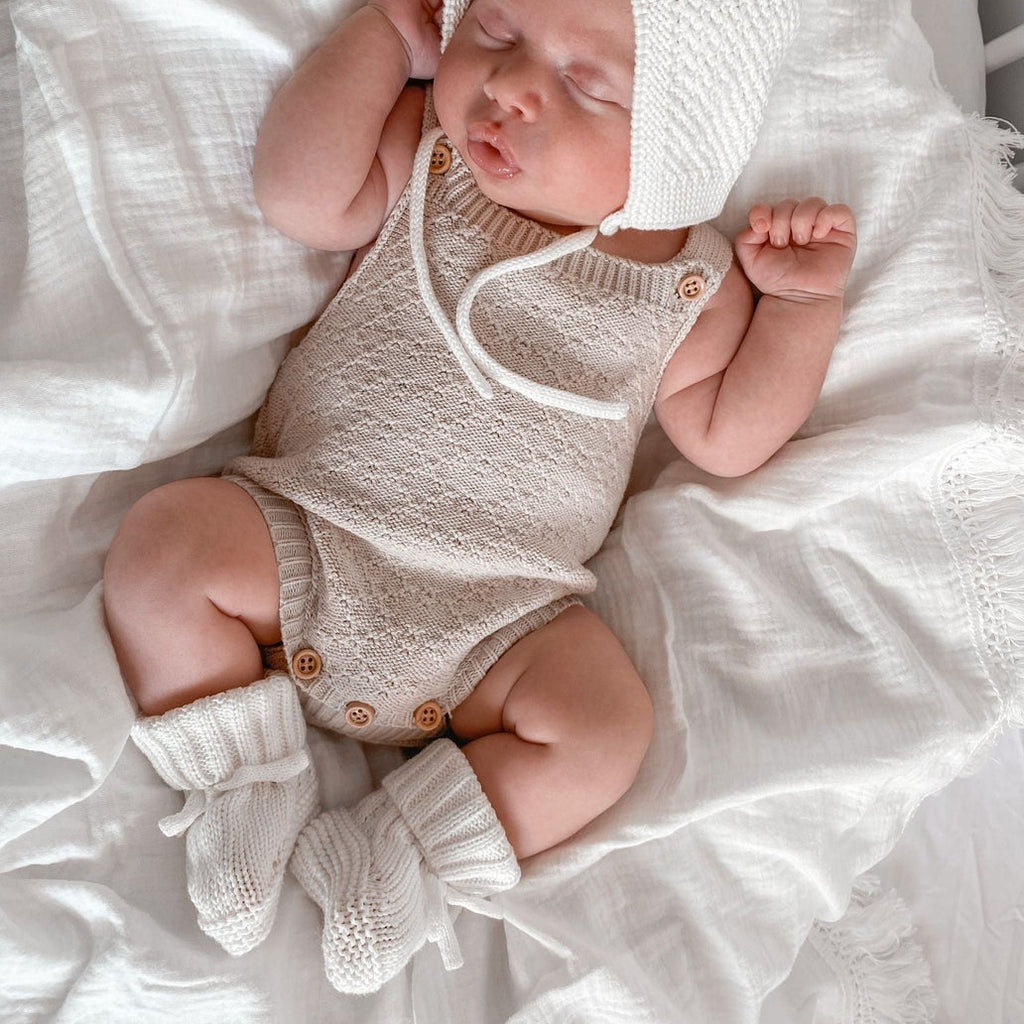 Newborn baby in a knitted outfit and bonnet lying on white bedding.