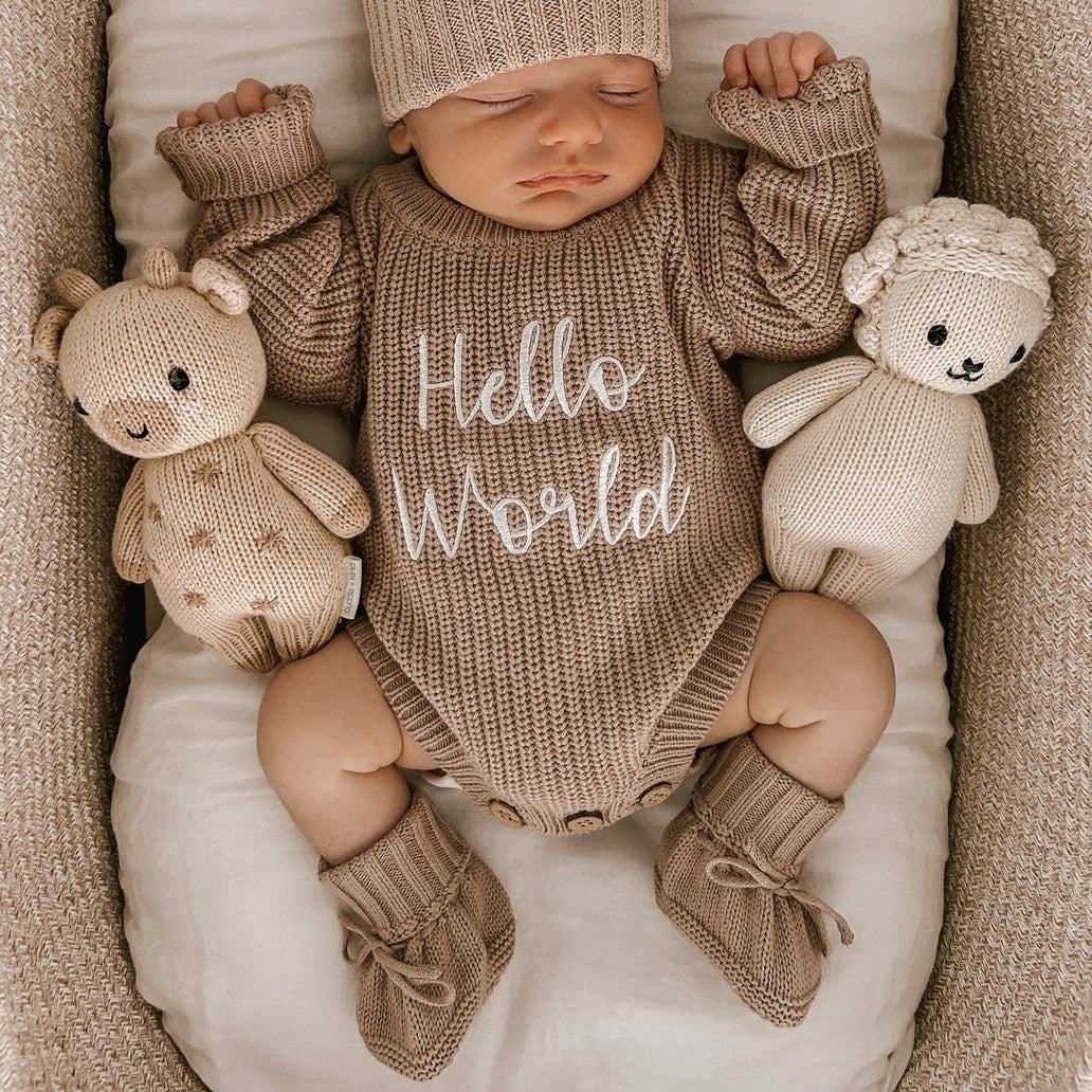 Newborn baby in a knitted outfit with 'Hello World' text, surrounded by teddy bears in a crib.