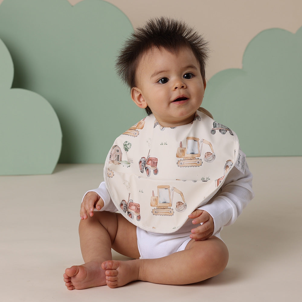Baby sitting on a beige surface wearing a bib with diggers and tractors designs against a green and beige cloud-patterned wall.