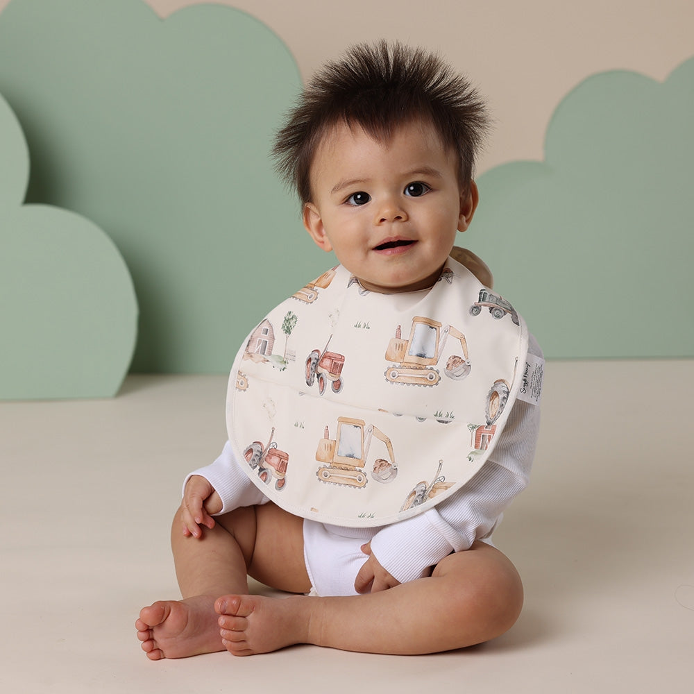 Baby wearing a bib with digger designs against a green and beige cloud-shaped backdrop