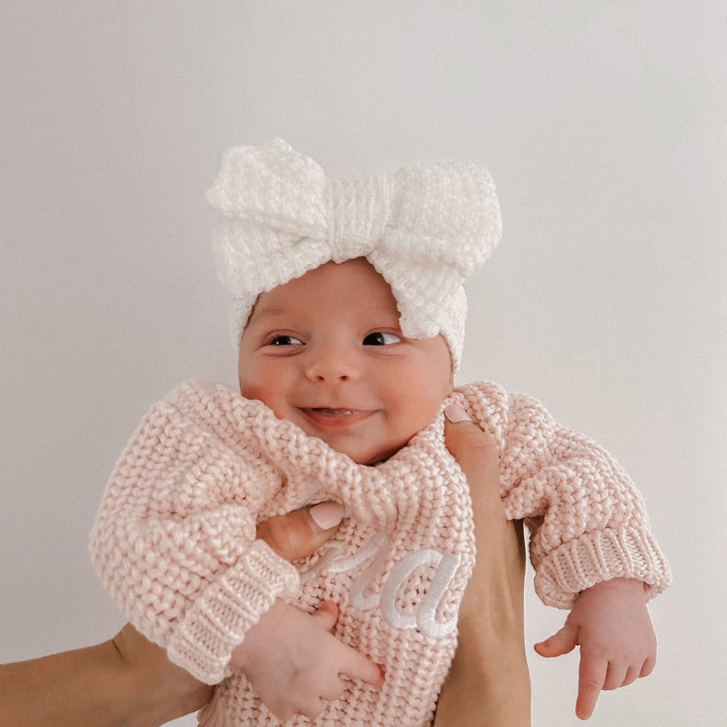 Baby wearing a pink knitted sweater and white bow headband, held by an adult against a plain background.