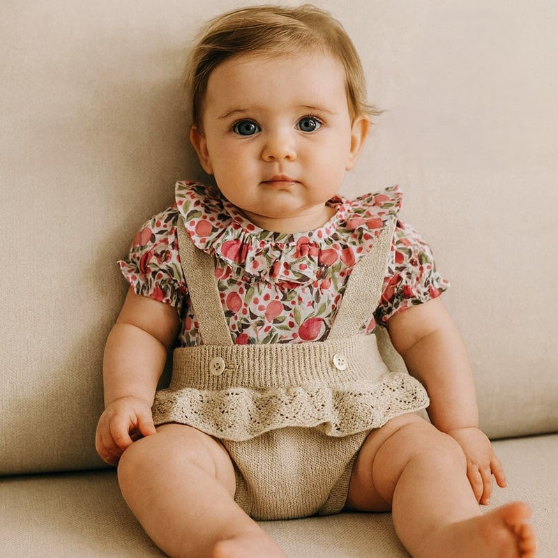 Baby wearing a floral top and beige overalls sitting on a beige couch.