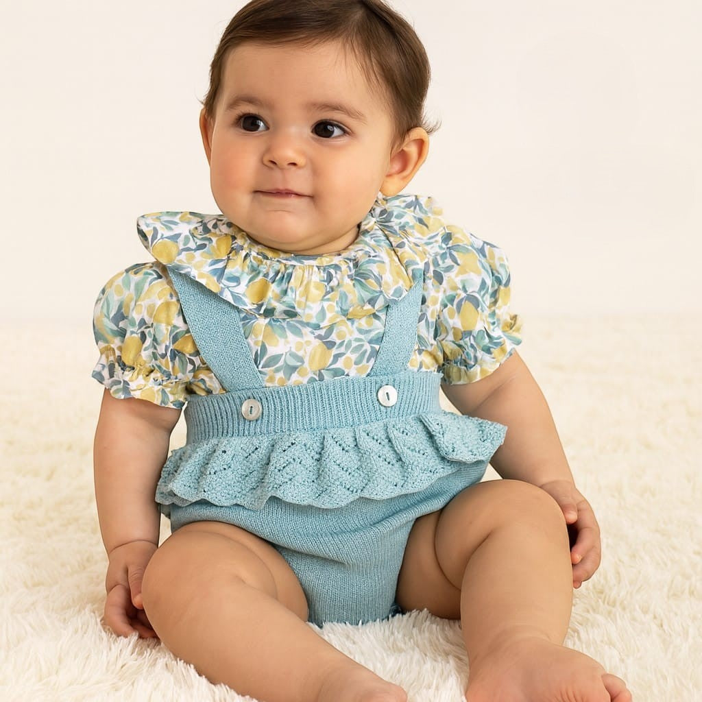 Baby wearing a floral top and blue ruffled overalls sitting on a white surface.
