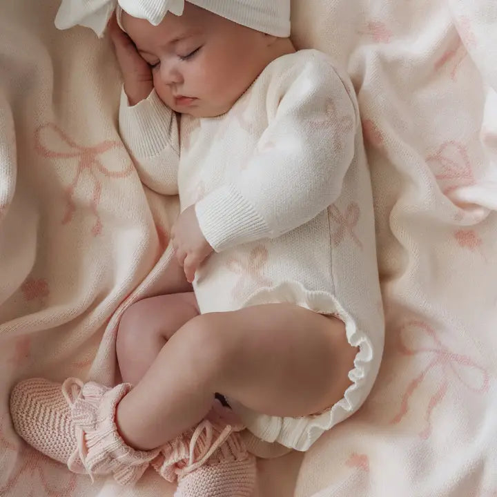 Baby in a white outfit and pink knitted booties lying on a soft pink blanket.