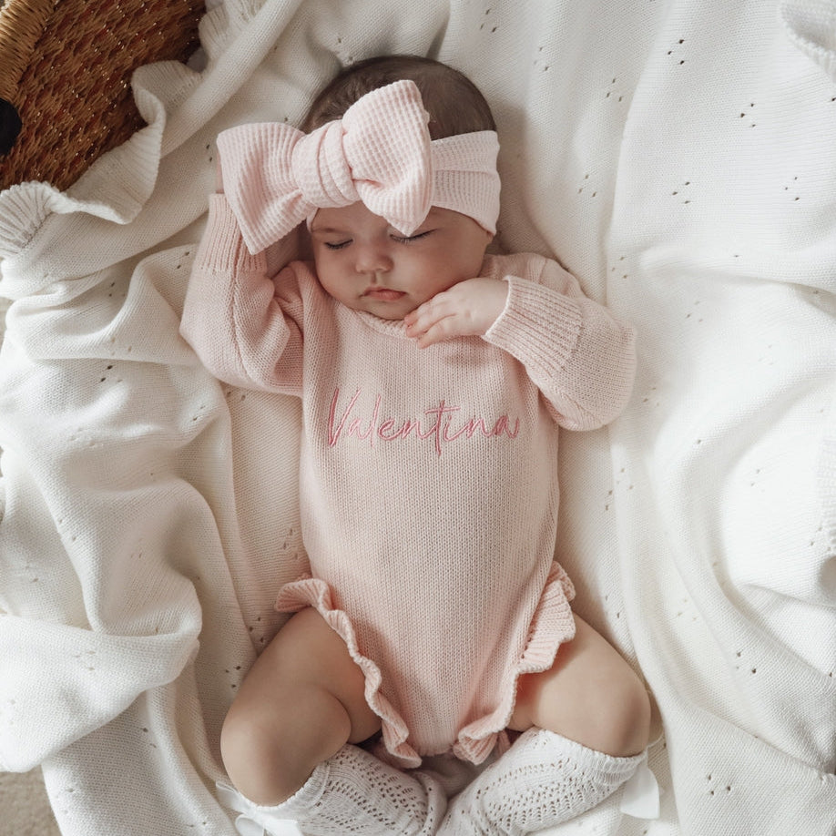 Newborn baby in pink outfit with large bow sitting on white blanket