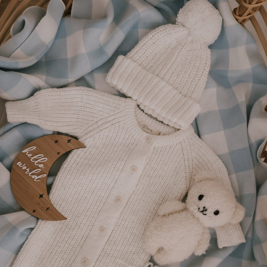 Knitted baby outfit with hat, mittens, and teddy bear toy on a checkered blanket.
