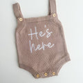 Knitted baby romper with 'He's here' text on a light gray background