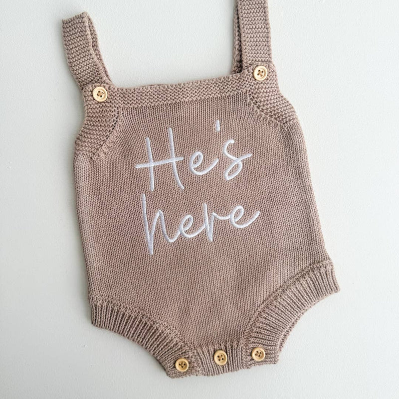 Knitted baby romper with 'He's here' text on a light gray background