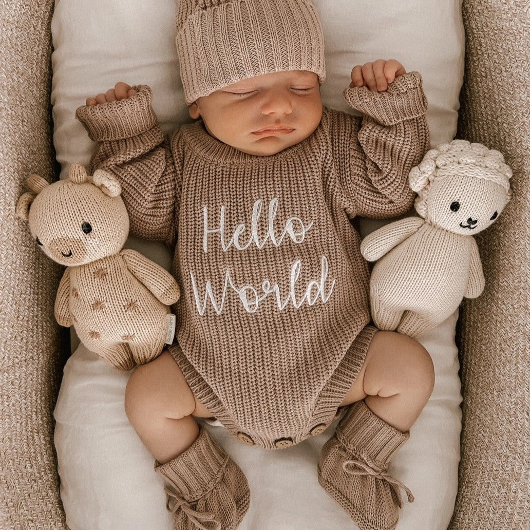 Newborn baby in a 'Hello World' outfit with knitted toys in a crib.