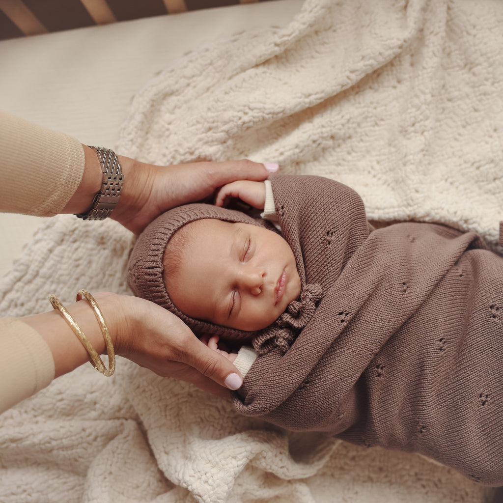 Newborn baby wrapped in brown swaddle with headband, lying on a soft surface.