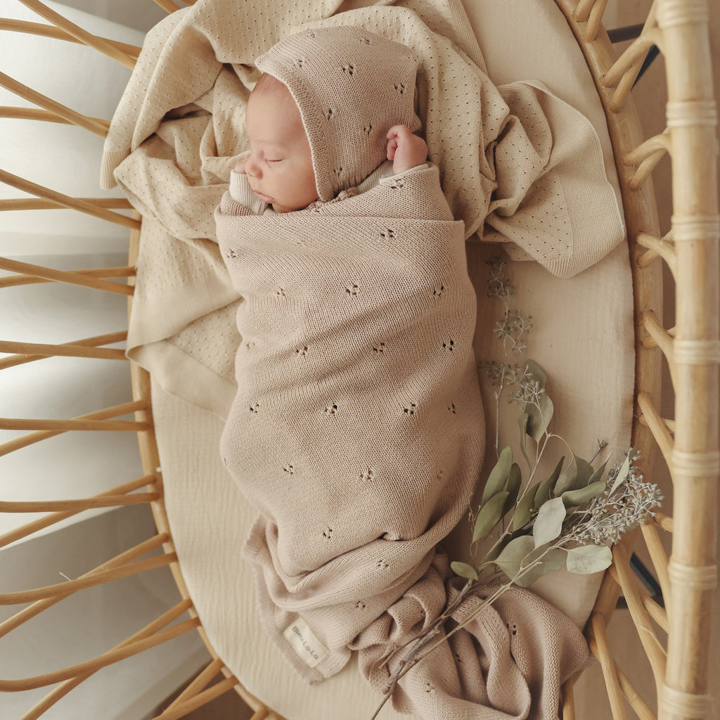 Newborn baby wrapped in a beige swaddle with a matching bonnet, sitting in a wicker basket.