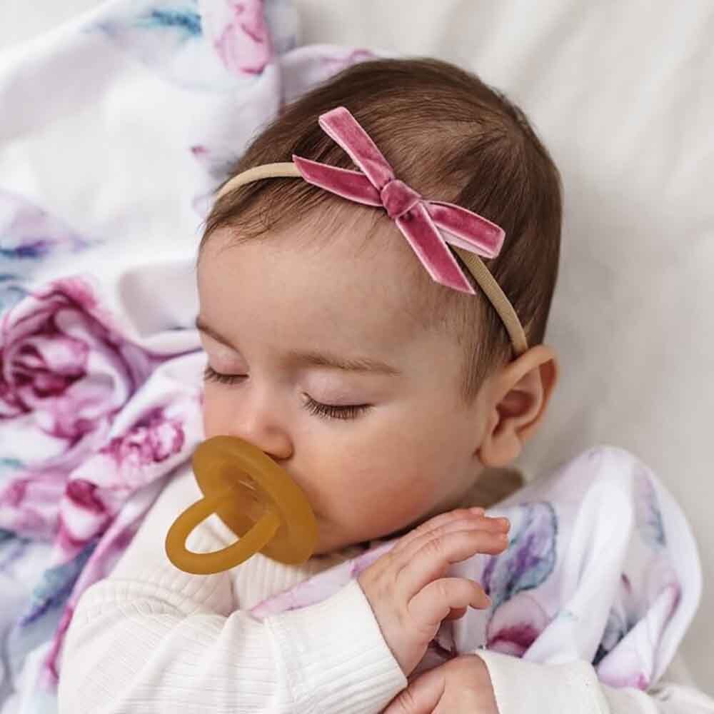 Baby with a pacifier and pink headband lying on a soft surface