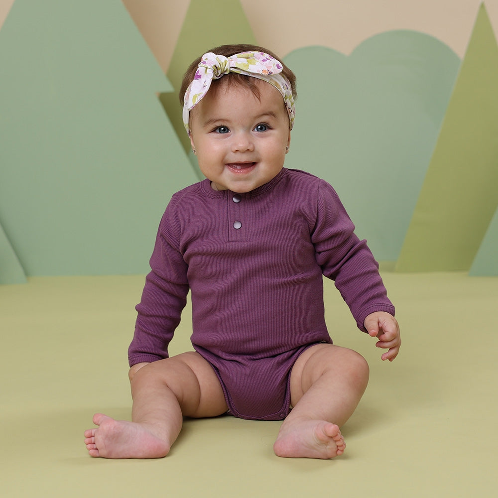 Baby wearing a purple onesie with a floral headband against a green and beige geometric-patterned background