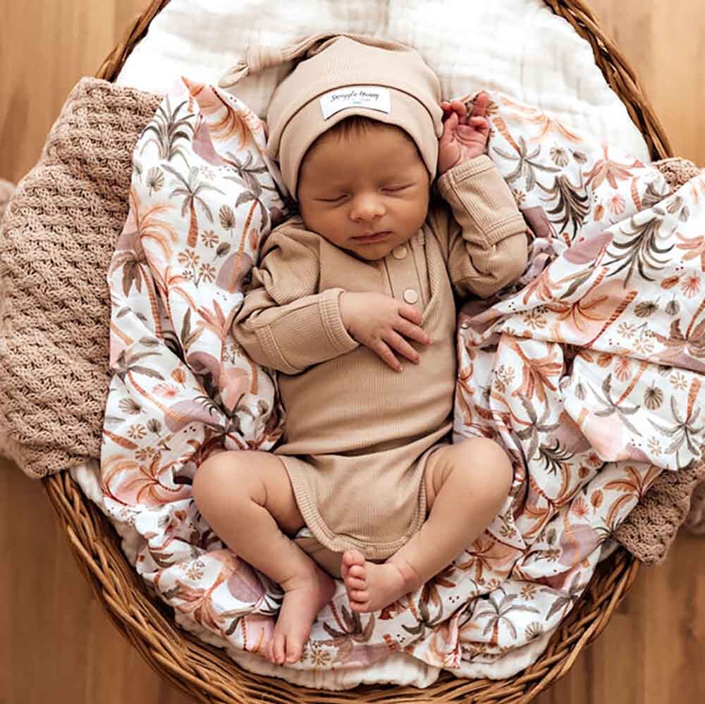 Newborn baby in a beige outfit and hat lying on floral blankets in a wicker basket.