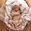 Newborn baby in a beige outfit and hat lying on floral blankets in a wicker basket.