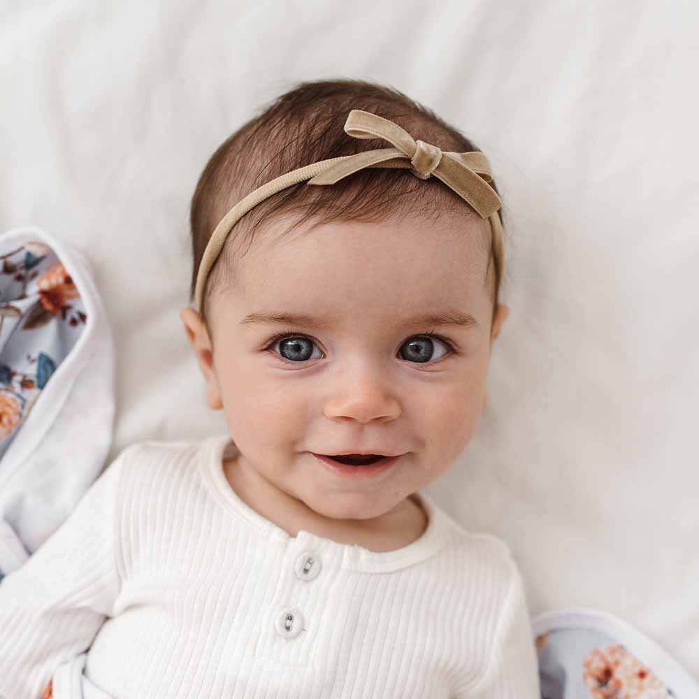 Baby wearing a beige headband with a bow, lying on a white blanket.