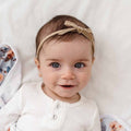 Baby wearing a beige headband with a bow, lying on a white blanket.