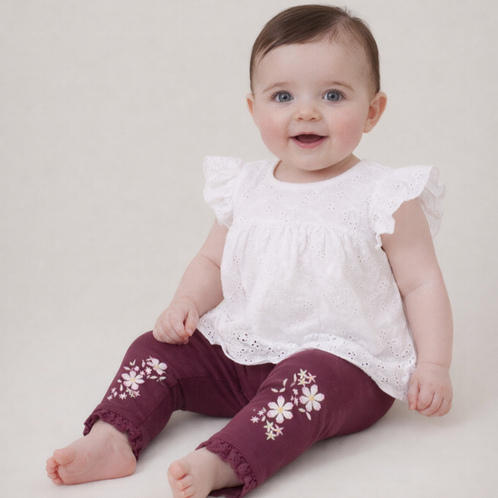 Baby wearing a white top and maroon pants with floral patterns on a plain background