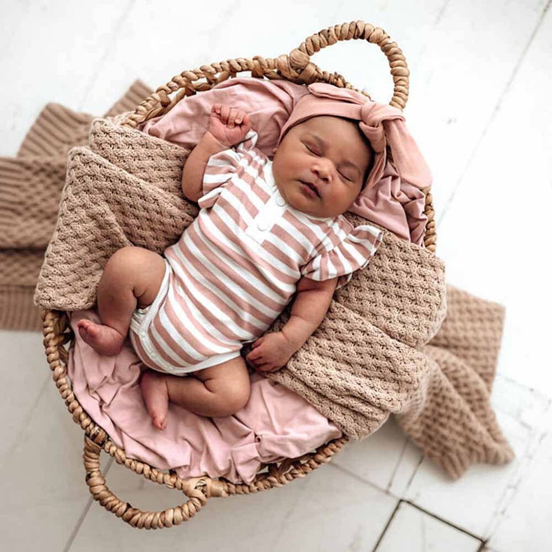 Newborn baby in a striped outfit and pink headband lying in a woven basket with pink blanket and beige knitted blanket.