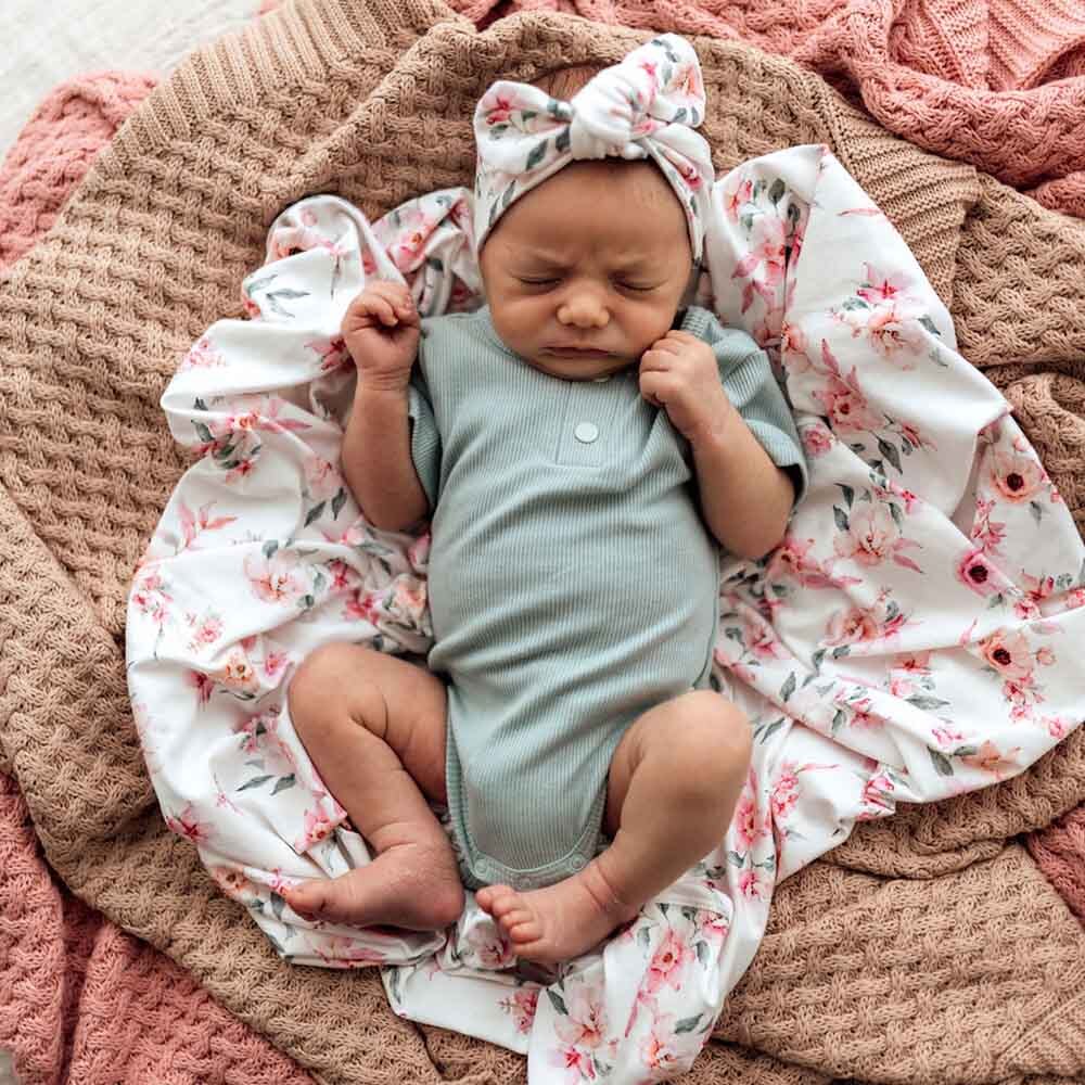 Newborn baby in a light green onesie with a floral headband and blanket on a pink textured surface