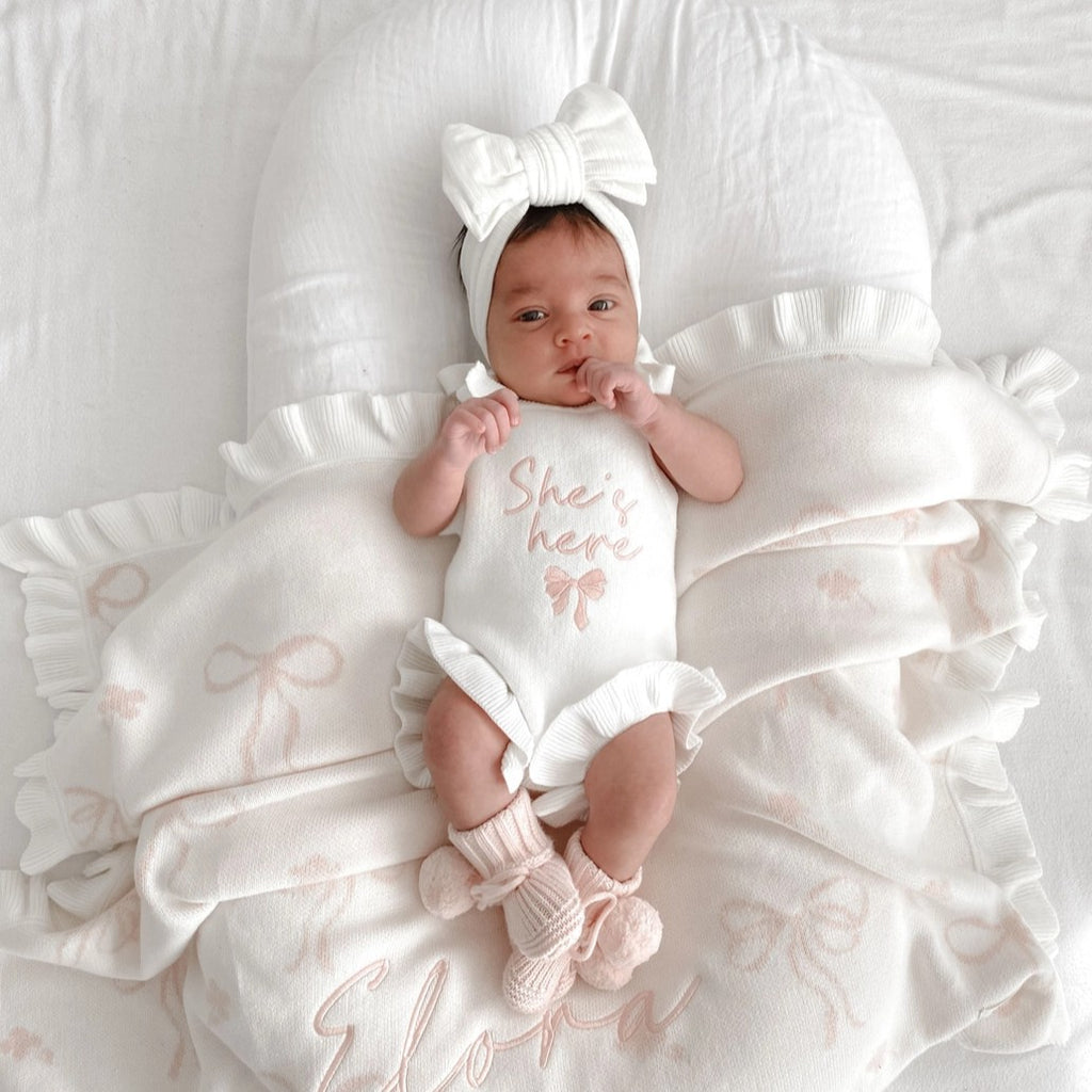 Baby in a white outfit with a bow and 'She is here' text, lying on a white textured surface.