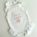White baby romper with 'She's here' embroidery on a light gray background
