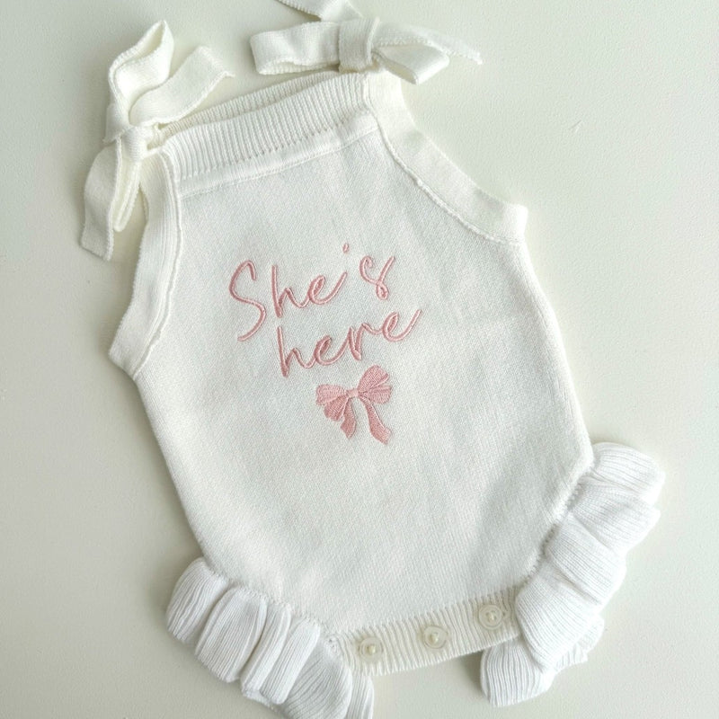 White baby romper with 'She's here' embroidery on a light gray background