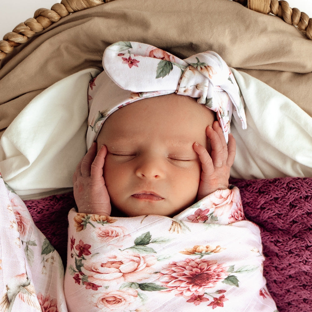 Newborn baby swaddled in floral blanket with matching headband, lying on textured surface.