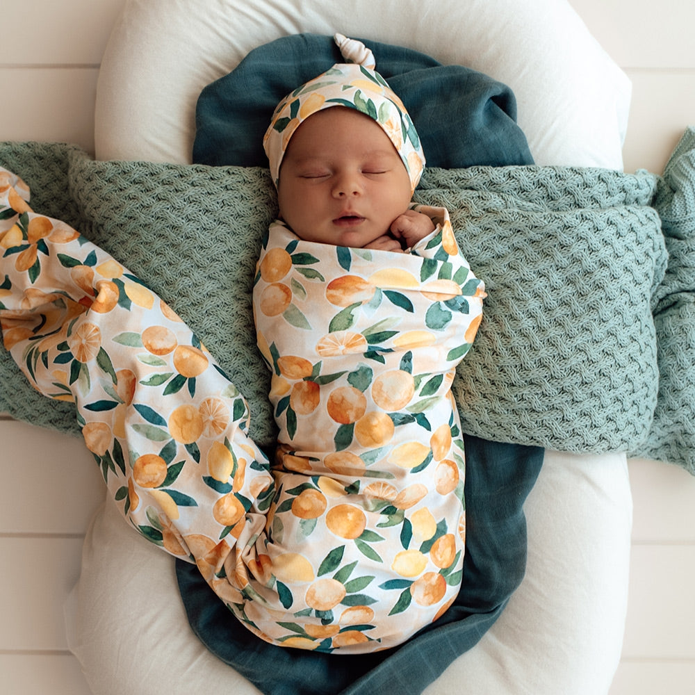 Newborn baby swaddled in Snuggle Hunny citrus printed wrap with matching knot beanie from Meridian Baby.