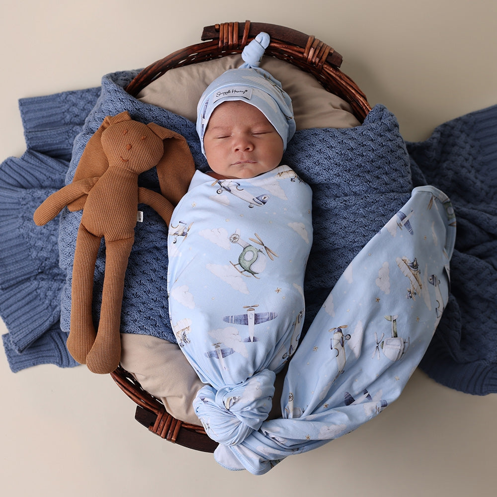 Newborn baby wrapped in a blue blanket with airplane pattern, wearing a matching hat, lying in a basket with a brown stuffed toy.
