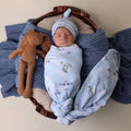 Newborn baby wrapped in a blue blanket with airplane pattern, wearing a matching hat, lying in a basket with a brown stuffed toy.
