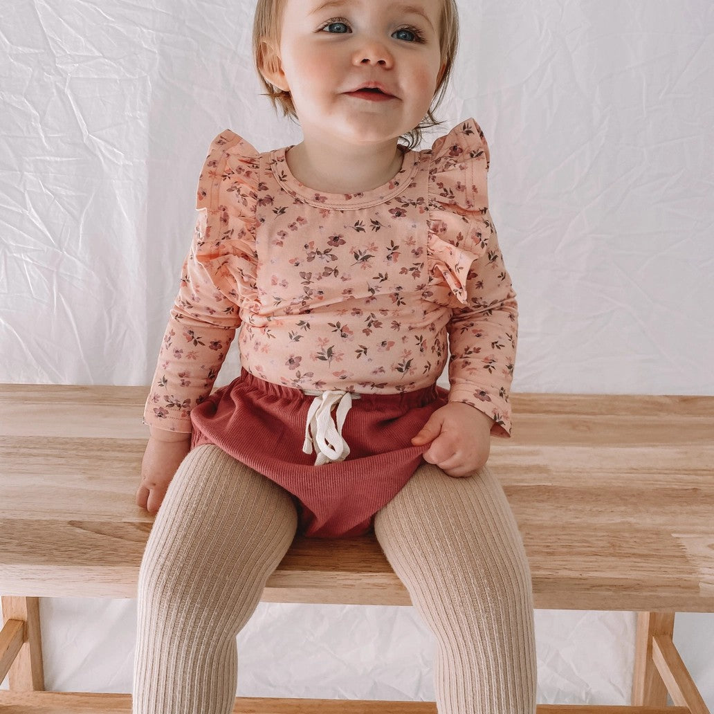 Child wearing a floral top and red shorts and beige tights sitting on a wooden bench.